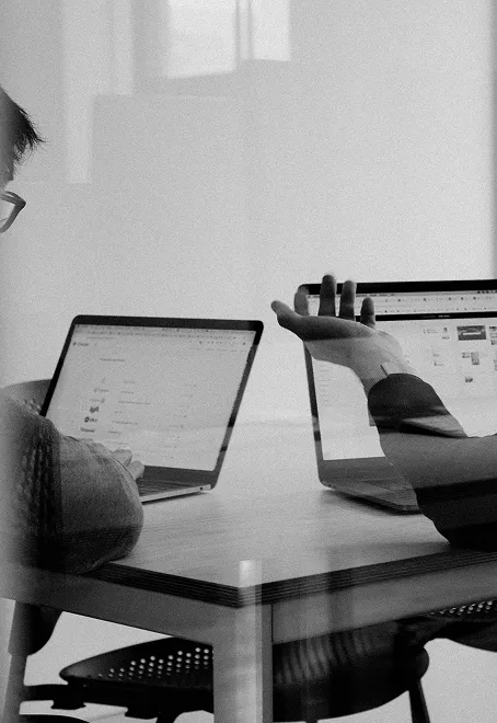 Person working at desk with laptop and external monitor, holding phone, black and white photograph showing dual screen workspace setup