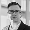 Black and white portrait of a person wearing glasses and dark clothing, photographed in professional lighting with a clean, minimalist backdrop