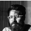 Black and white portrait of a bearded man wearing glasses, photographed against a curtained background in dramatic lighting